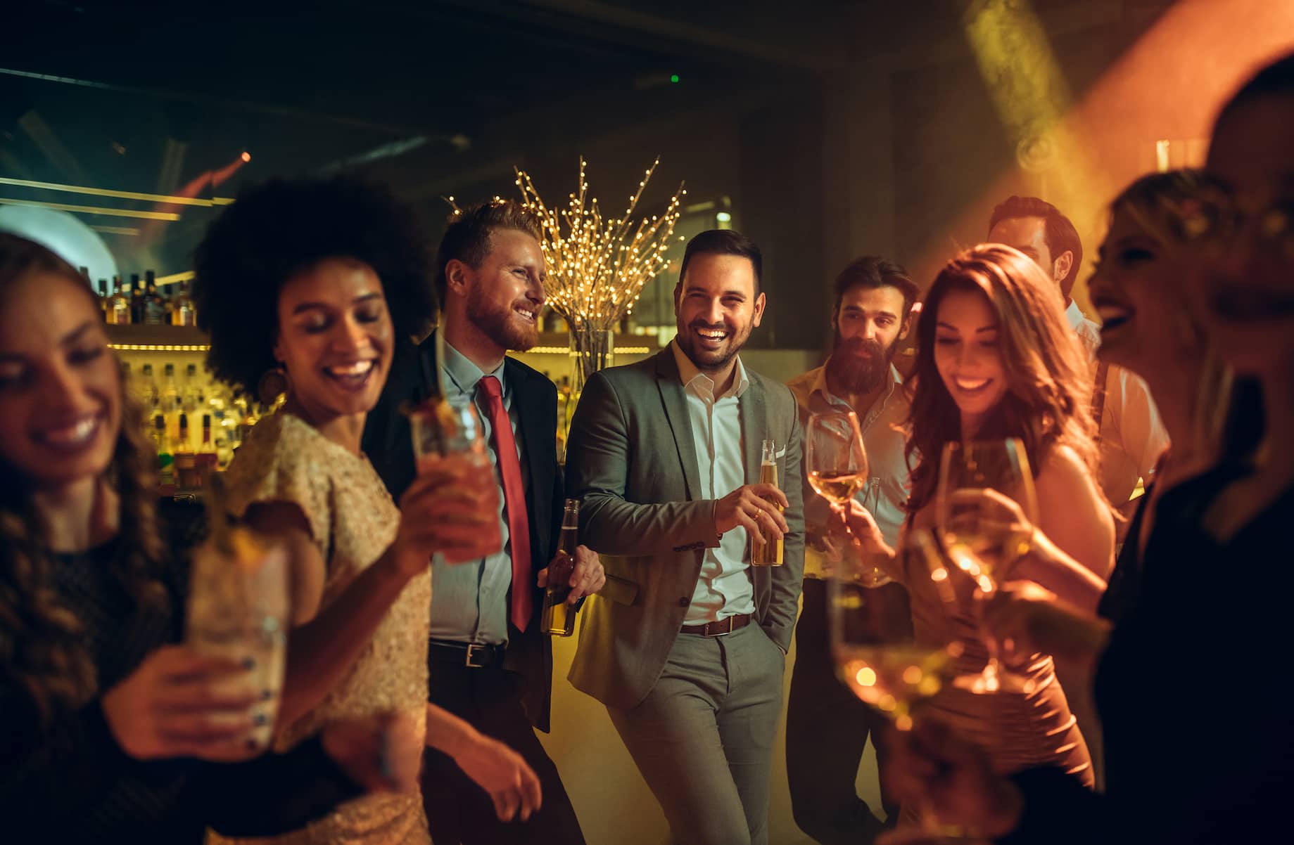 A group of smiling people holding drinks, enjoying a lively celebration in a bar setting with festive lighting.