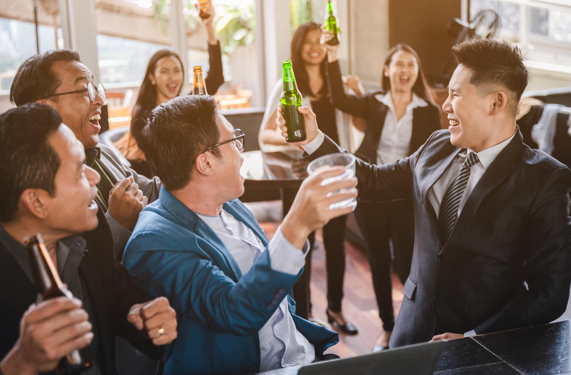 A group of professionals celebrating with drinks, smiling and raising glasses in a lively indoor setting.