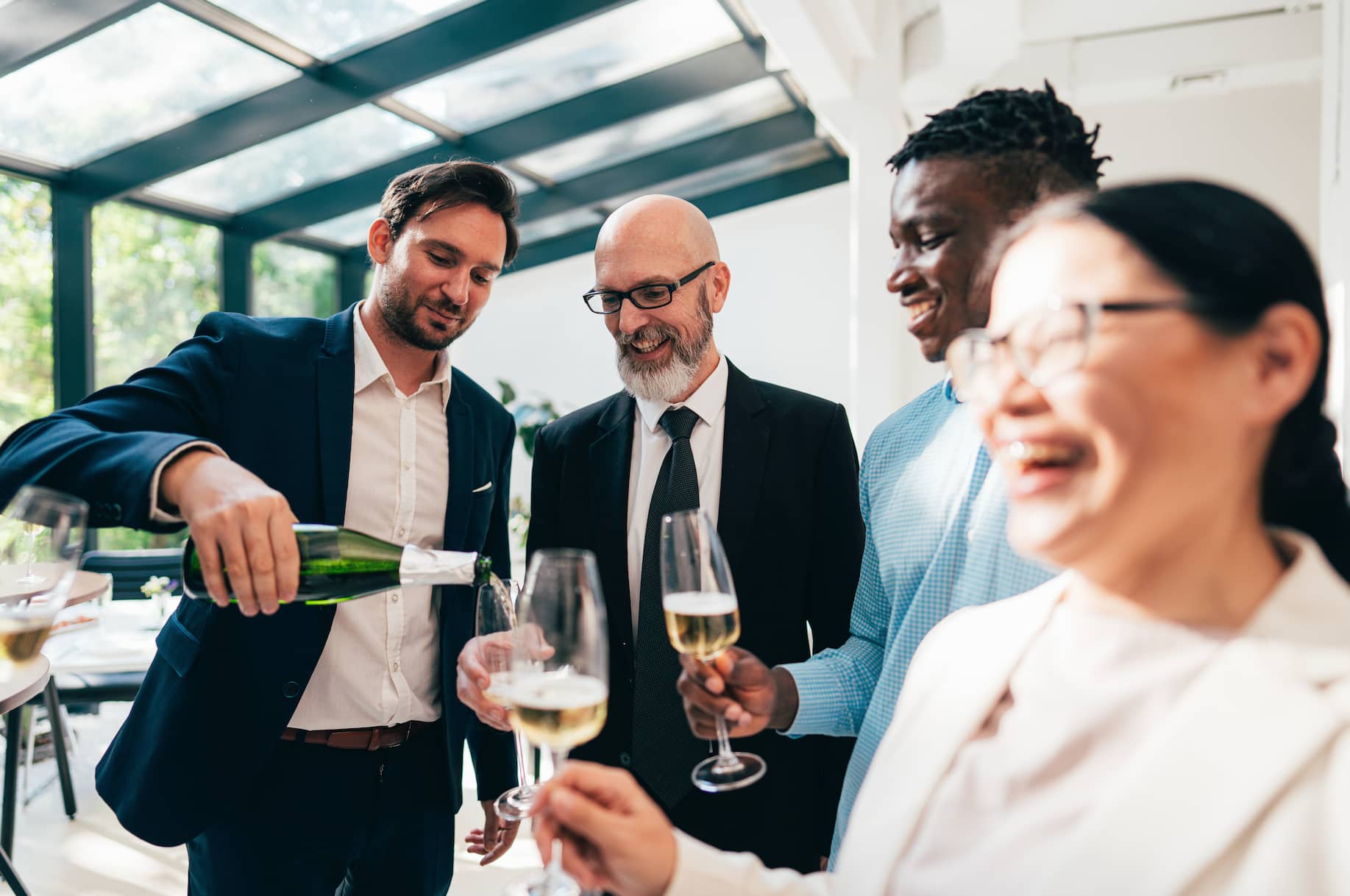 A group of four people is toasting with champagne glasses, with one person pouring champagne from a bottle. They are dressed in formal attire and are smiling in a well-lit indoor setting.