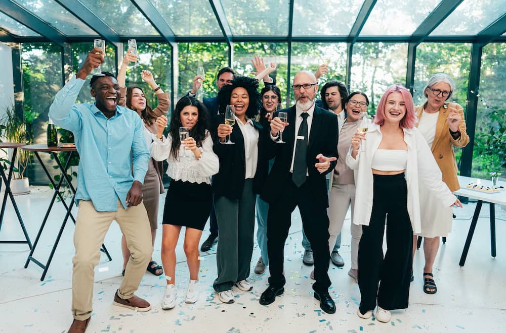 A group of diverse people celebrating together, holding drinks and smiling in a bright, festive indoor setting.