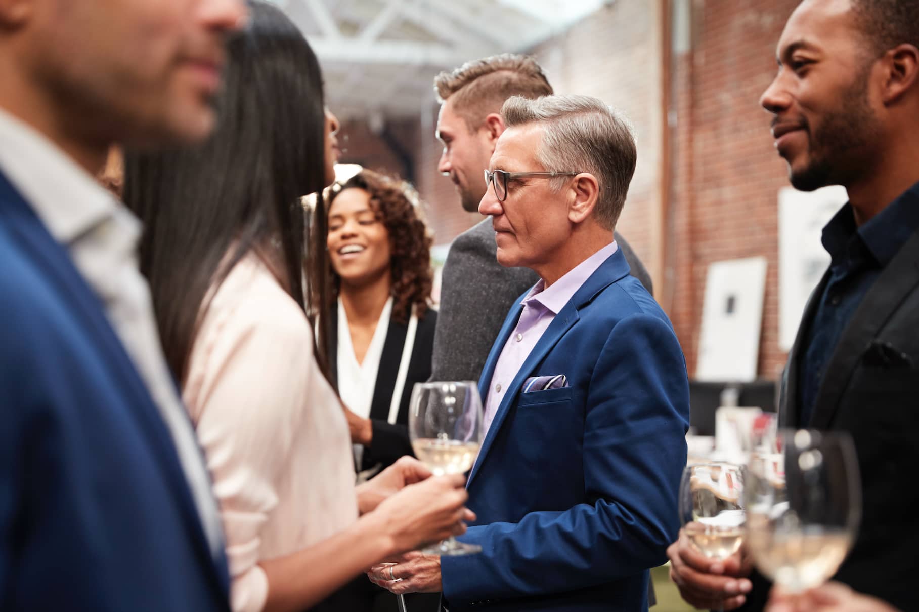 A group of people socializing at an event, some holding glasses of wine, with a focus on a man in a blue suit and glasses.