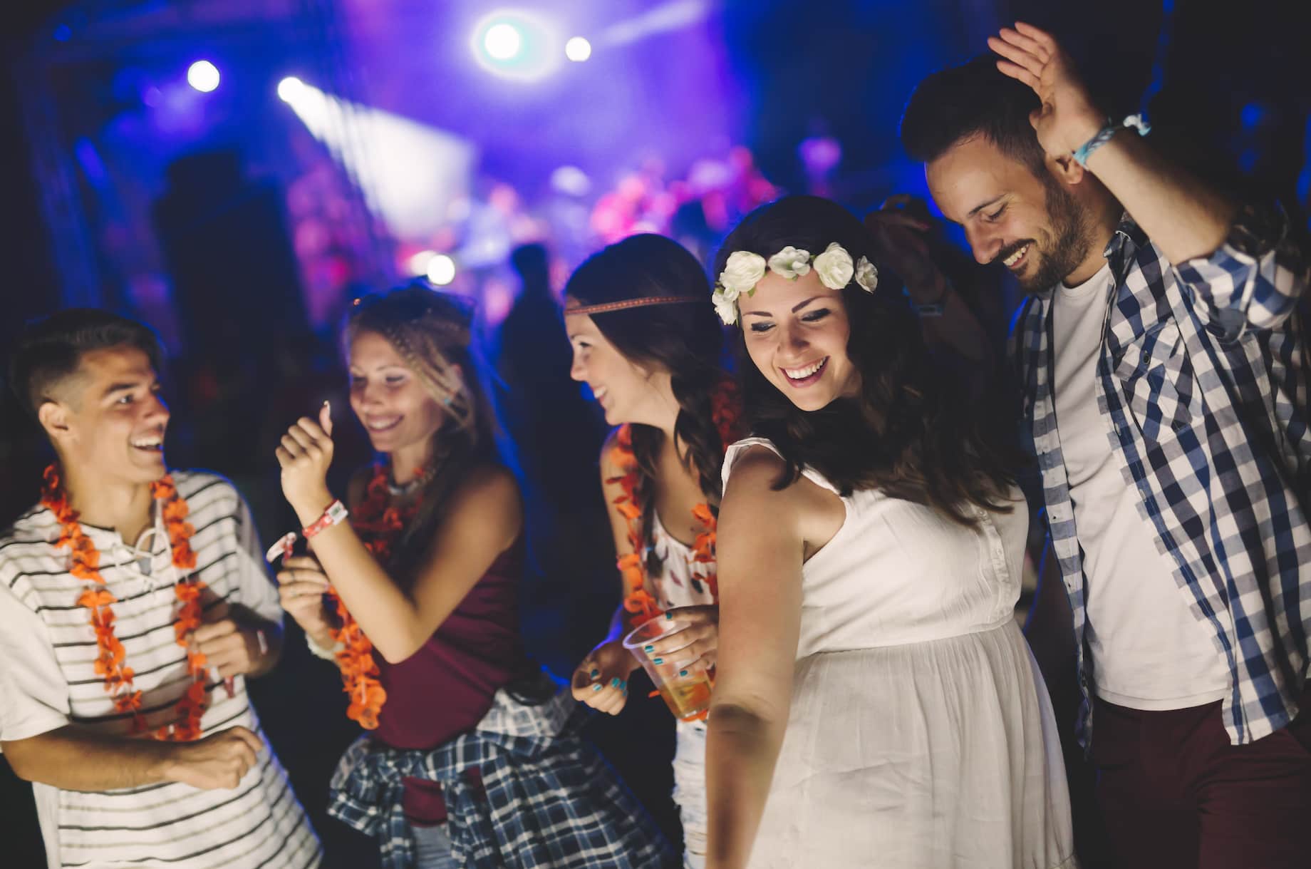A group of cheerful young adults dancing at a lively event, wearing colorful leis and enjoying drinks in a vibrant, illuminated atmosphere.