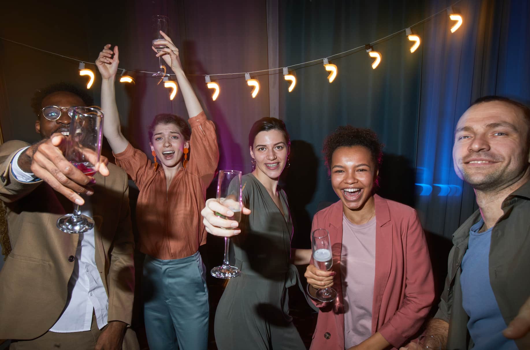A group of five people celebrating in a dimly lit setting, holding drinks and smiling, with festive lighting in the background.