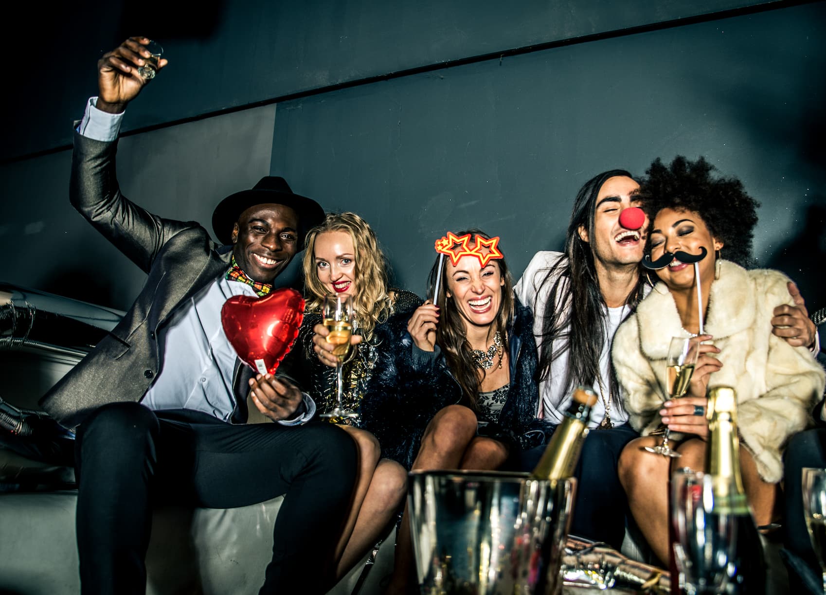 A group of five friends is celebrating at a party, smiling and posing with drinks. They wear festive accessories, including a heart-shaped balloon, star-shaped glasses, and a fake mustache. Champagne bottles and glasses are visible in the foreground.