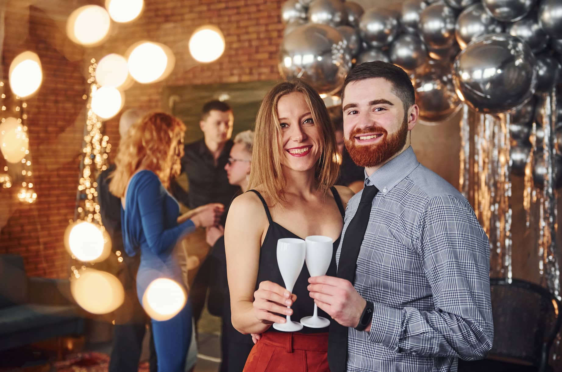A couple is smiling and holding white champagne flutes at a festive event, with blurred lights and guests in the background.
