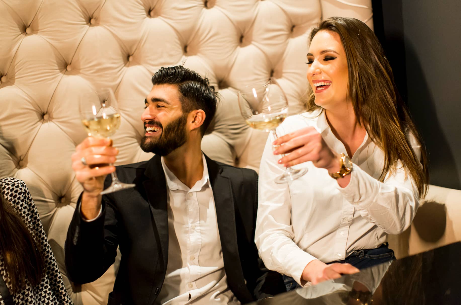 A man and a woman sit together, smiling and toasting with glasses of wine in a cozy setting.