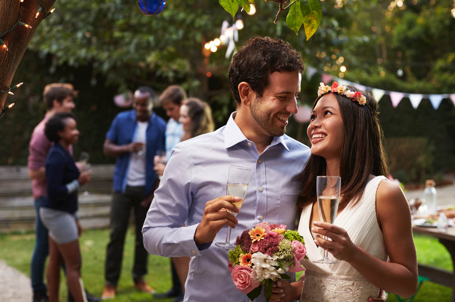 A couple is smiling at each other while holding champagne glasses and a bouquet of flowers at a festive outdoor gathering. Other guests are visible in the background.