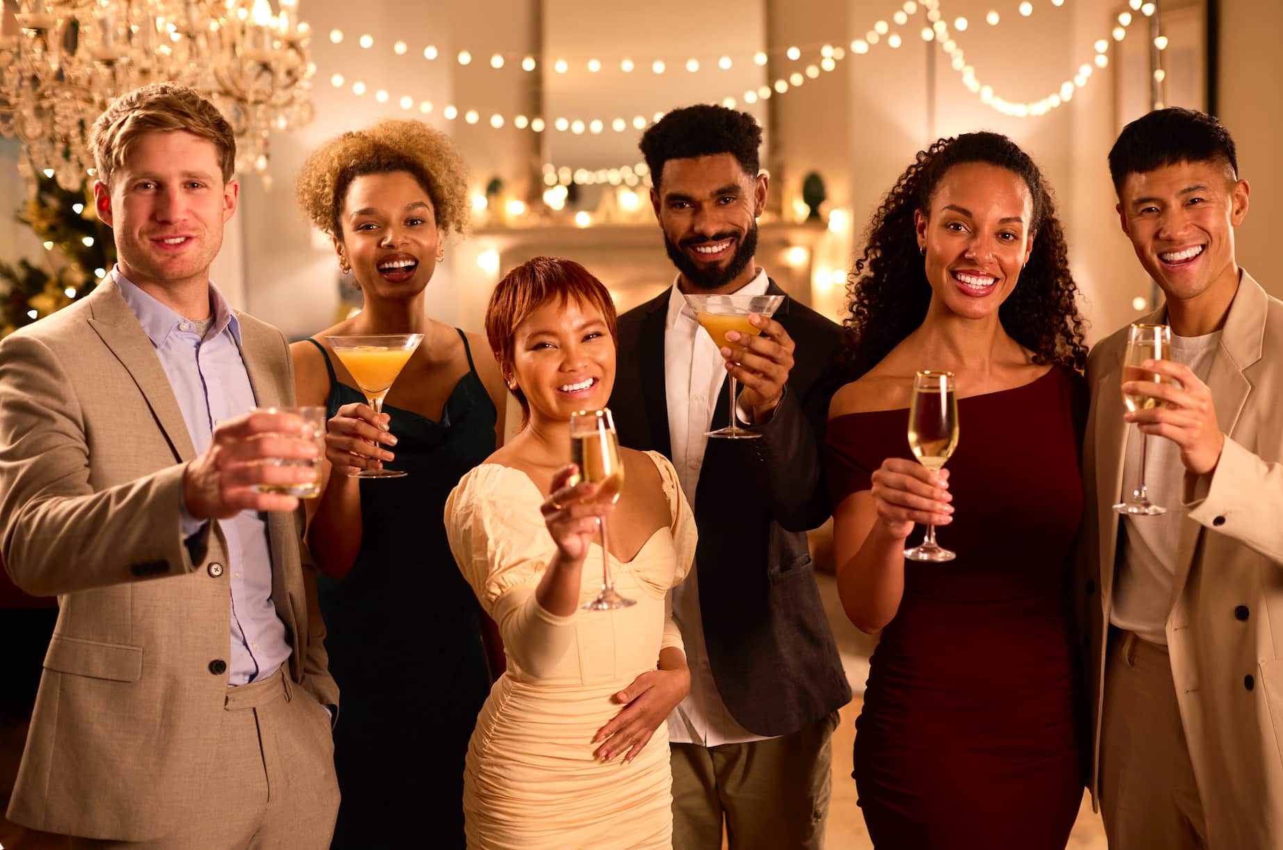 A group of six people cheerfully raising their drinks at a festive gathering, with decorative lights in the background.