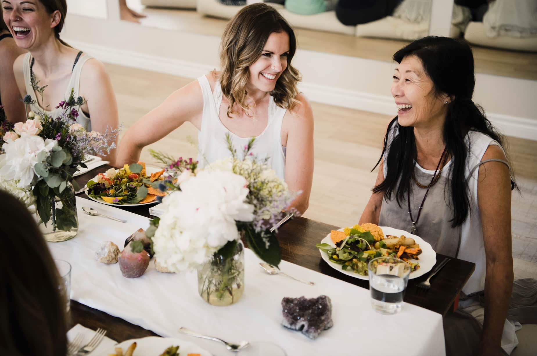 A group of women enjoying a meal together at a beautifully arranged table, featuring plates of colorful food and floral centerpieces. They are smiling and laughing, creating a warm, joyful atmosphere.