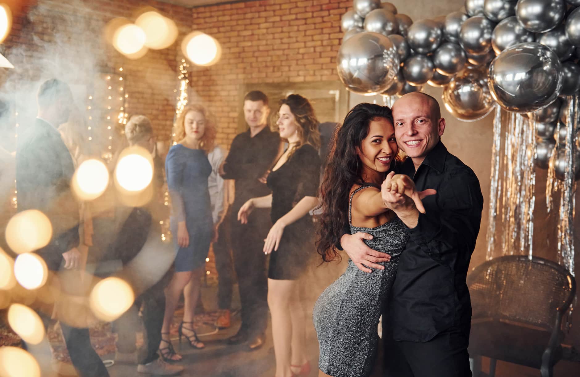 A couple dances joyfully in a festive setting with balloons and lights, while other guests socialize in the background.