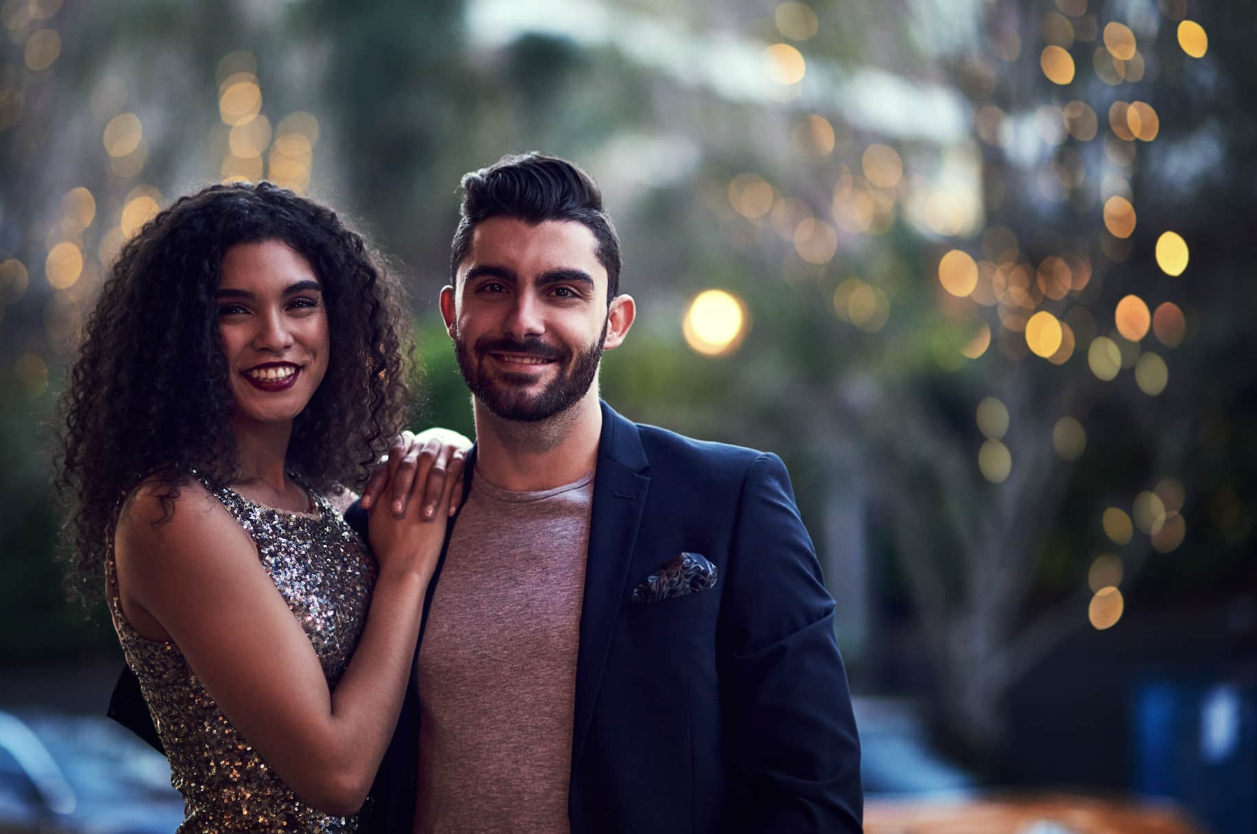 A woman in a sparkly dress and a man in a blazer are smiling together outdoors with a soft, blurred background featuring lights.