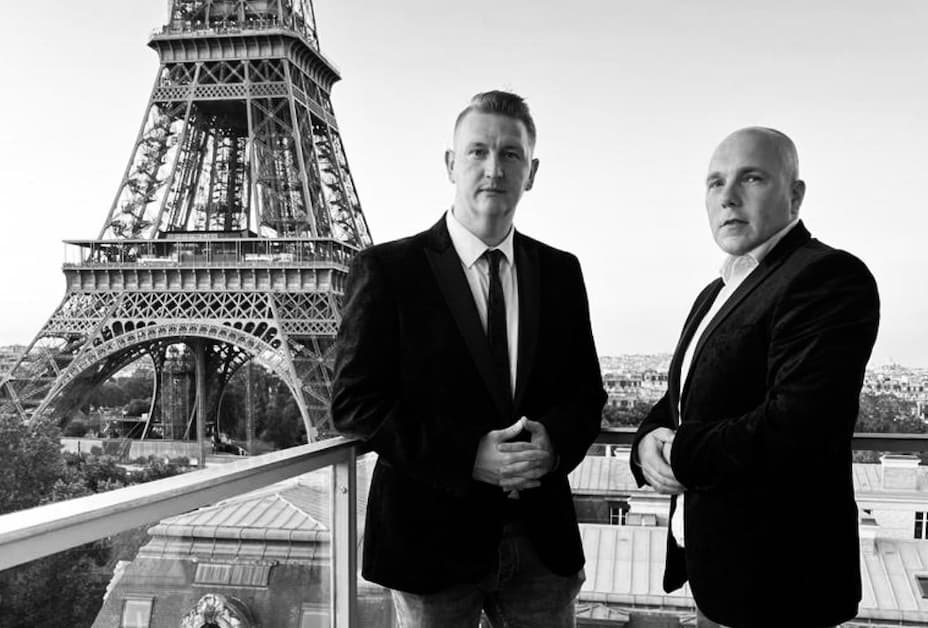 Two men in suits stand in front of the Eiffel Tower, which is visible in the background. The image is in black and white.