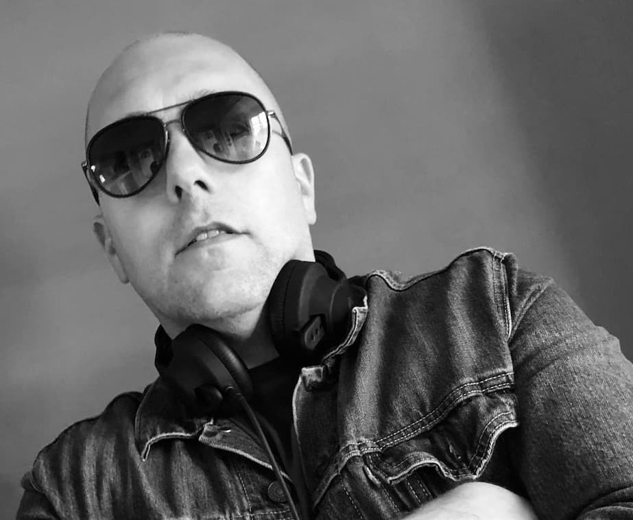 A person wearing sunglasses and headphones, posing with arms crossed, in a monochrome image.