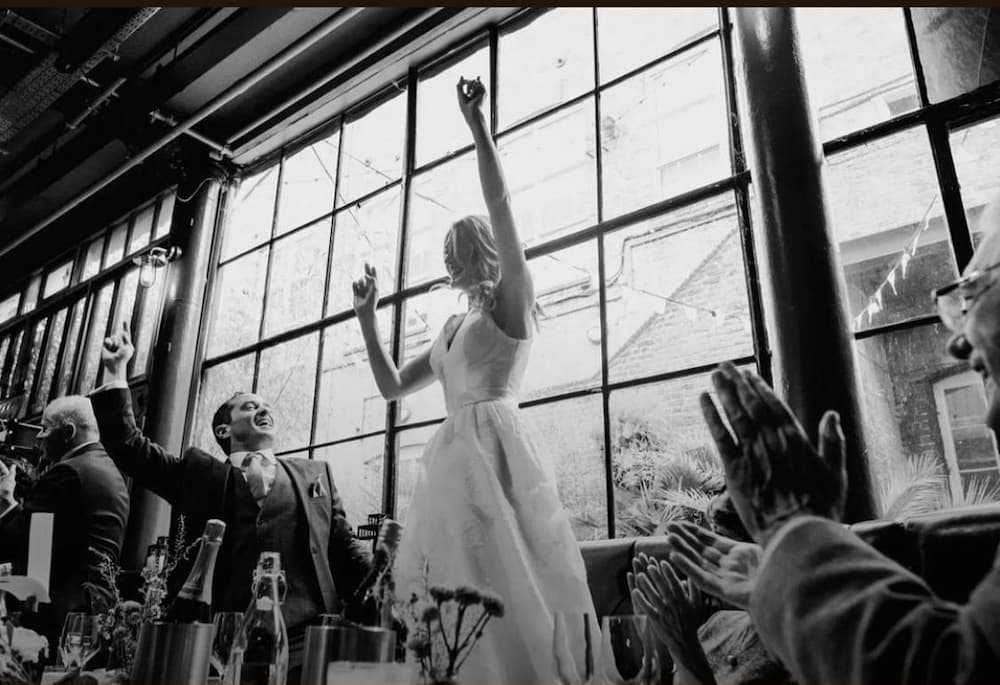 A woman in a white wedding dress is standing in front of a window, holding her hands up in the air. She is celebrating her wedding reception. The scene is set in a room with a dining table and several wine glasses placed on it.